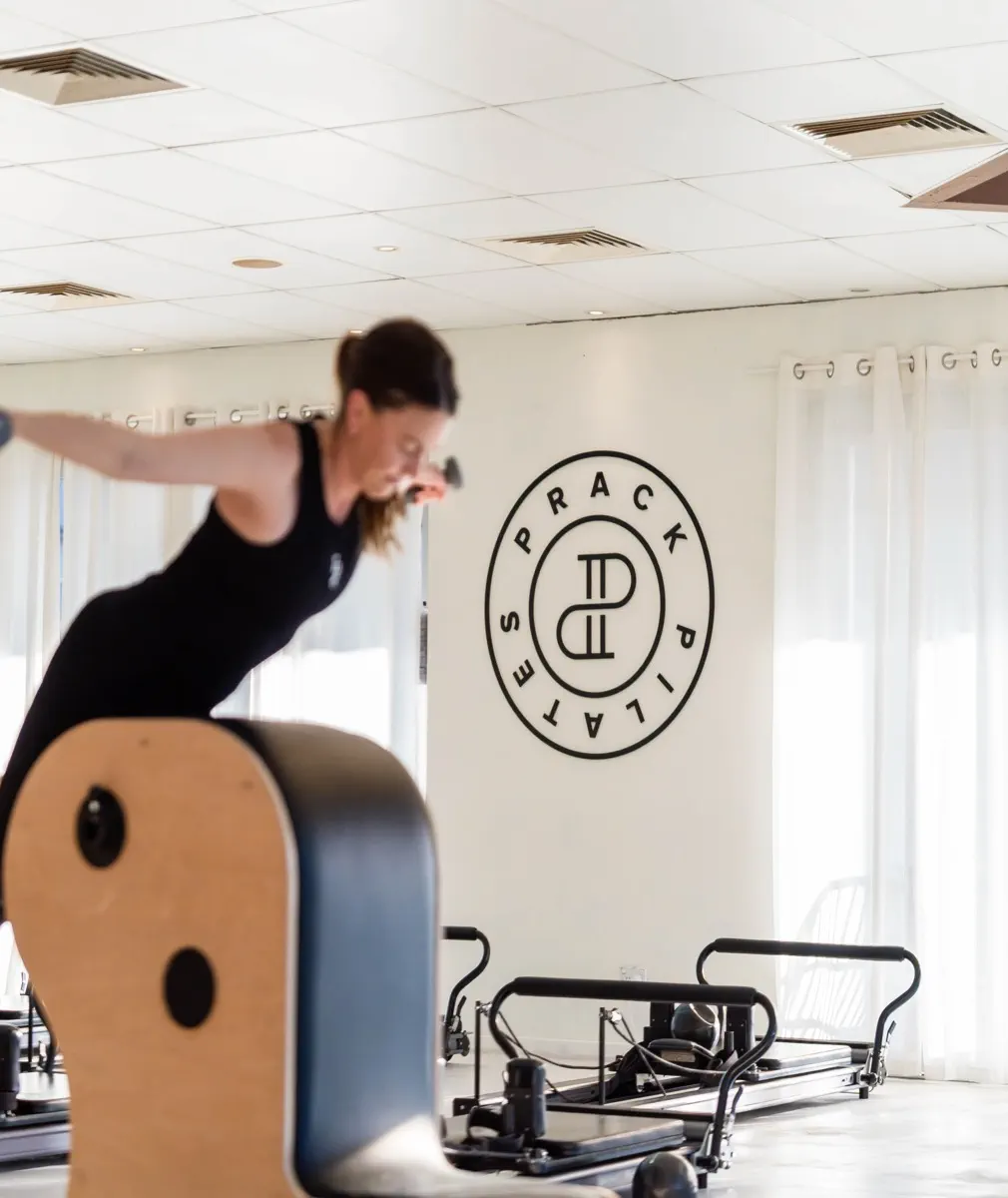 A client balanced over the Ladder Barrel holding weights, in front of the Prack Pilates roundel on the wall