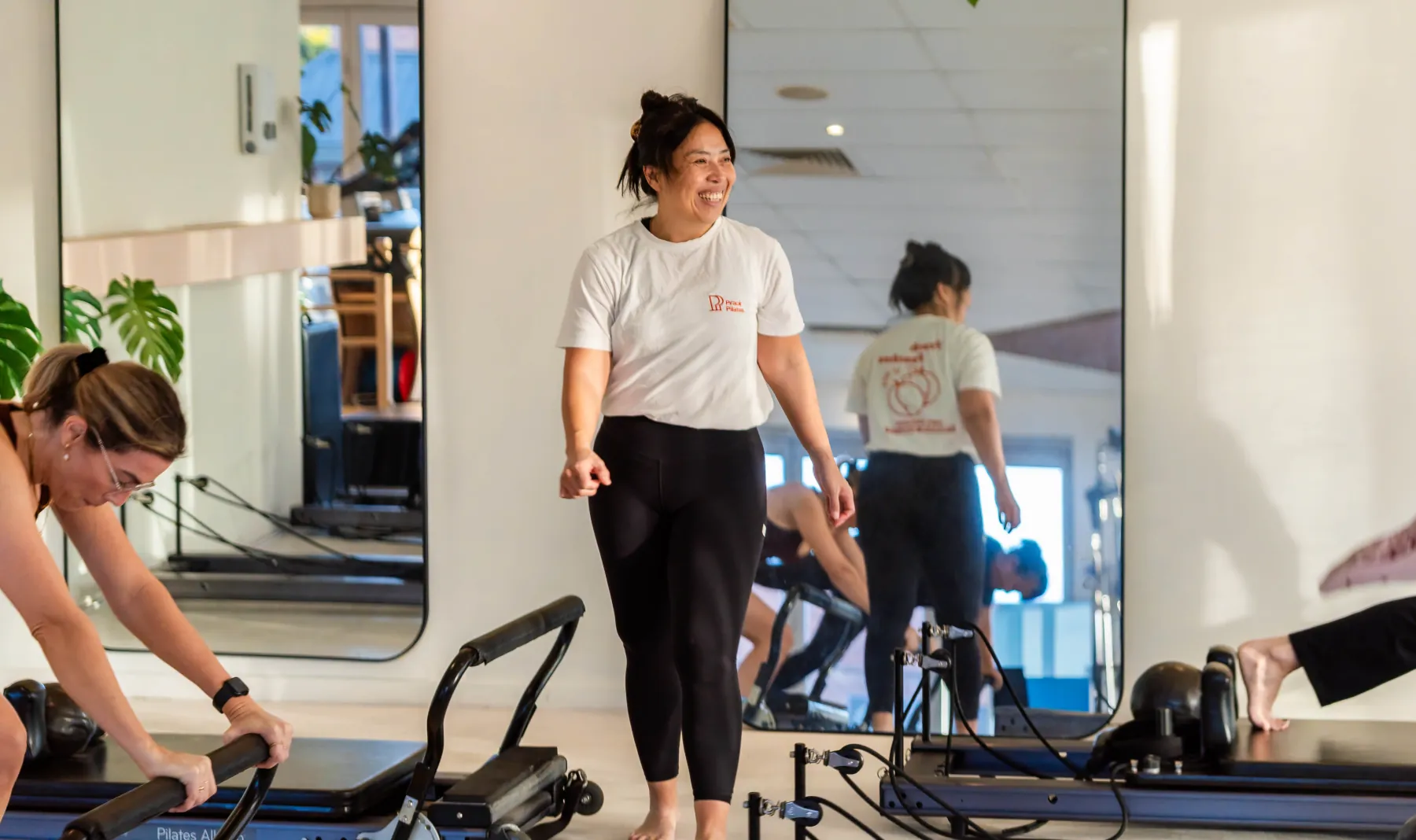 Souli in a Prack Pilates t-shirt, smiling while watching clients work on the reformers