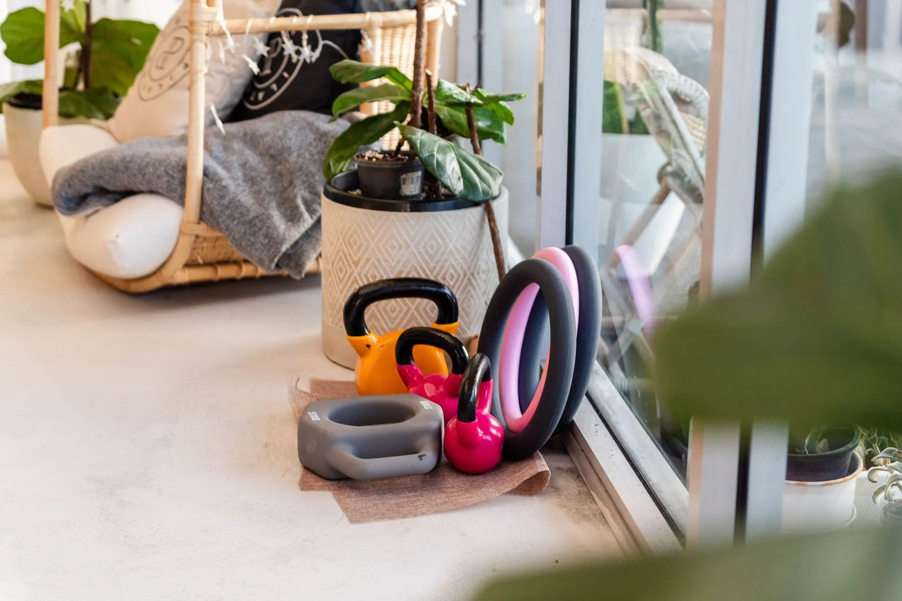 Kettlebells, Pilates rings and a weighted ring on the floor beside a rattan chair and plants.